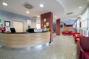 Una zona reception di un hotel con un banco di accoglienza curvo, sedute rosse e una sveglia appesa al muro.