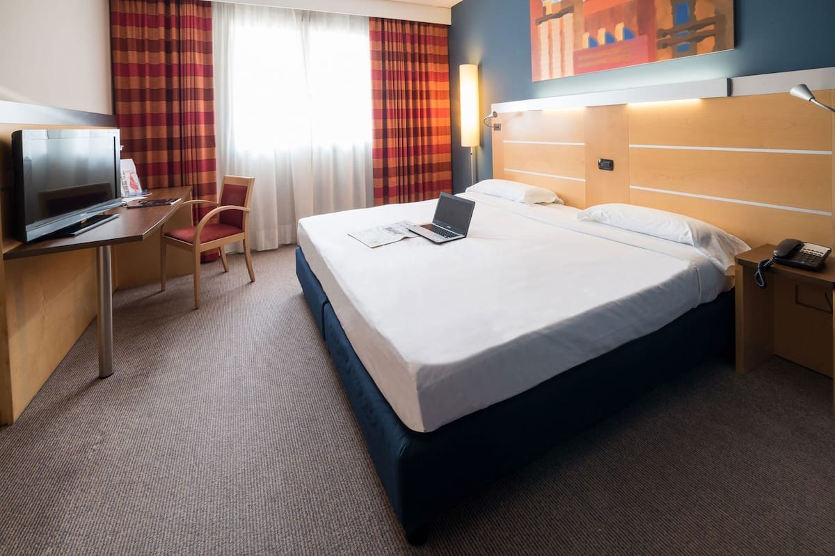 A hotel room with a bed, a desk with a laptop, a TV, a chair, and a bedside phone.