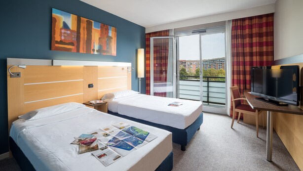 A hotel room with two beds, a desk with a TV, and a view of the city through the window. - Idea Hotel Torino Mirafiori (Turin)