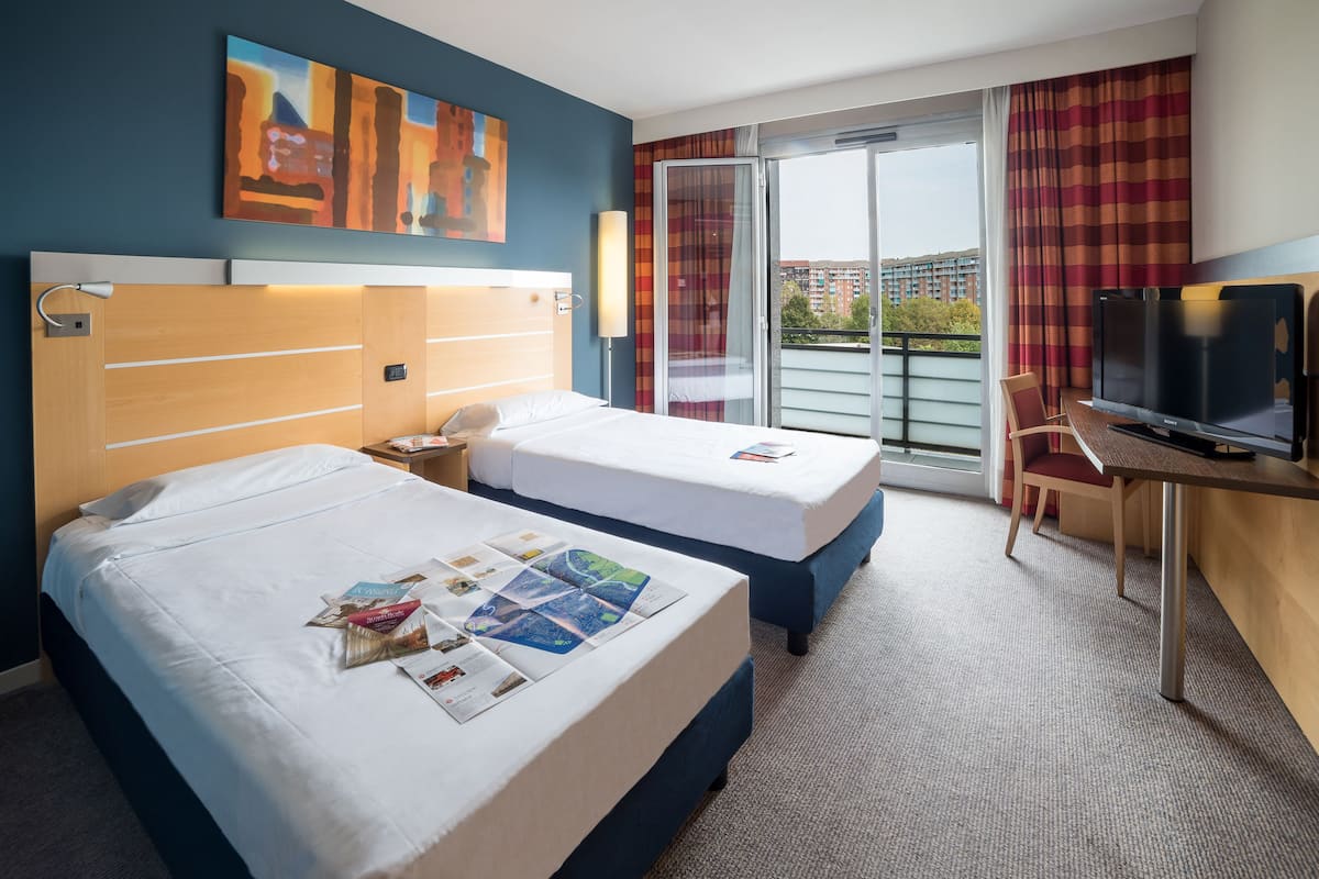 A hotel room with two beds, a desk with a TV, a large window with curtains, and a painting on the wall.