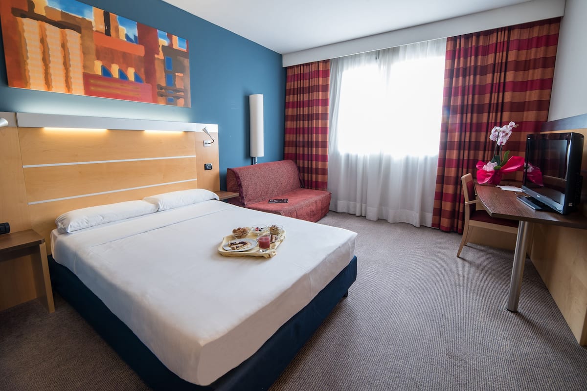 A hotel room with a bed, a desk with a TV, a chair, a painting on the wall, and a window with curtains.
