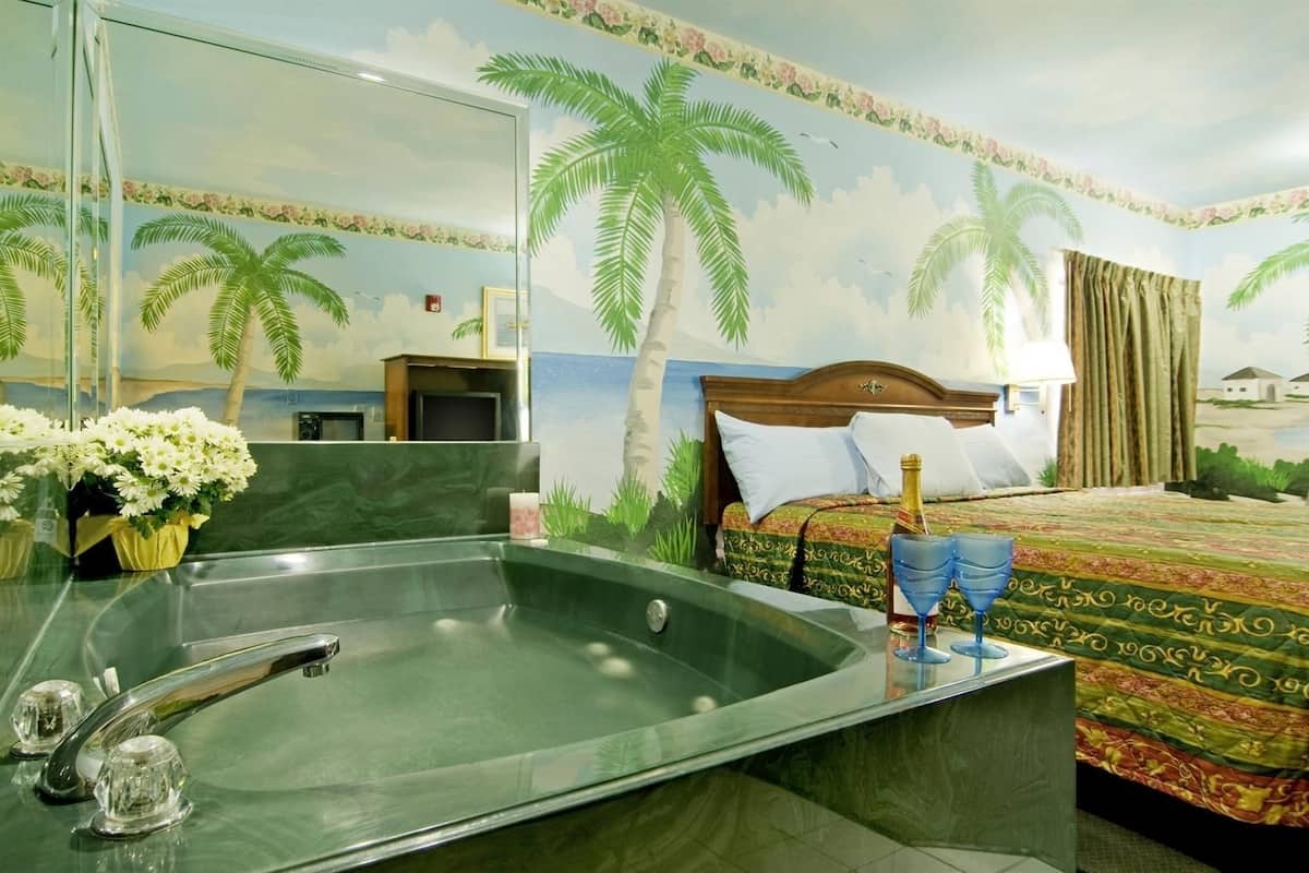 A hotel room with a large bathtub, a bed with a patterned bedspread, a wooden headboard, and a wall mural of palm trees.