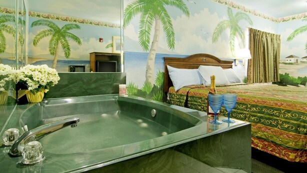 A hotel room with a large bathtub, a bed with a patterned bedspread, and a wall mural of palm trees. - Americas Best Value Inn Baytown (Baytown)