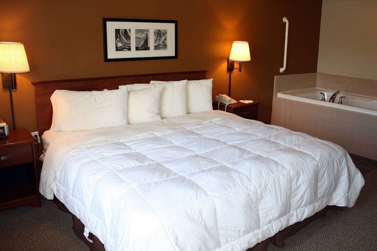 A hotel room with a large bed, bedside lamps, a small table with a phone, a wall-mounted picture, and a bathroom with a bathtub and sink.