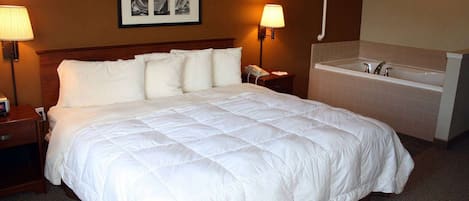 A hotel room with a large bed, bedside lamps, a small table with a phone, a wall-mounted picture, and a bathroom with a bathtub and sink.