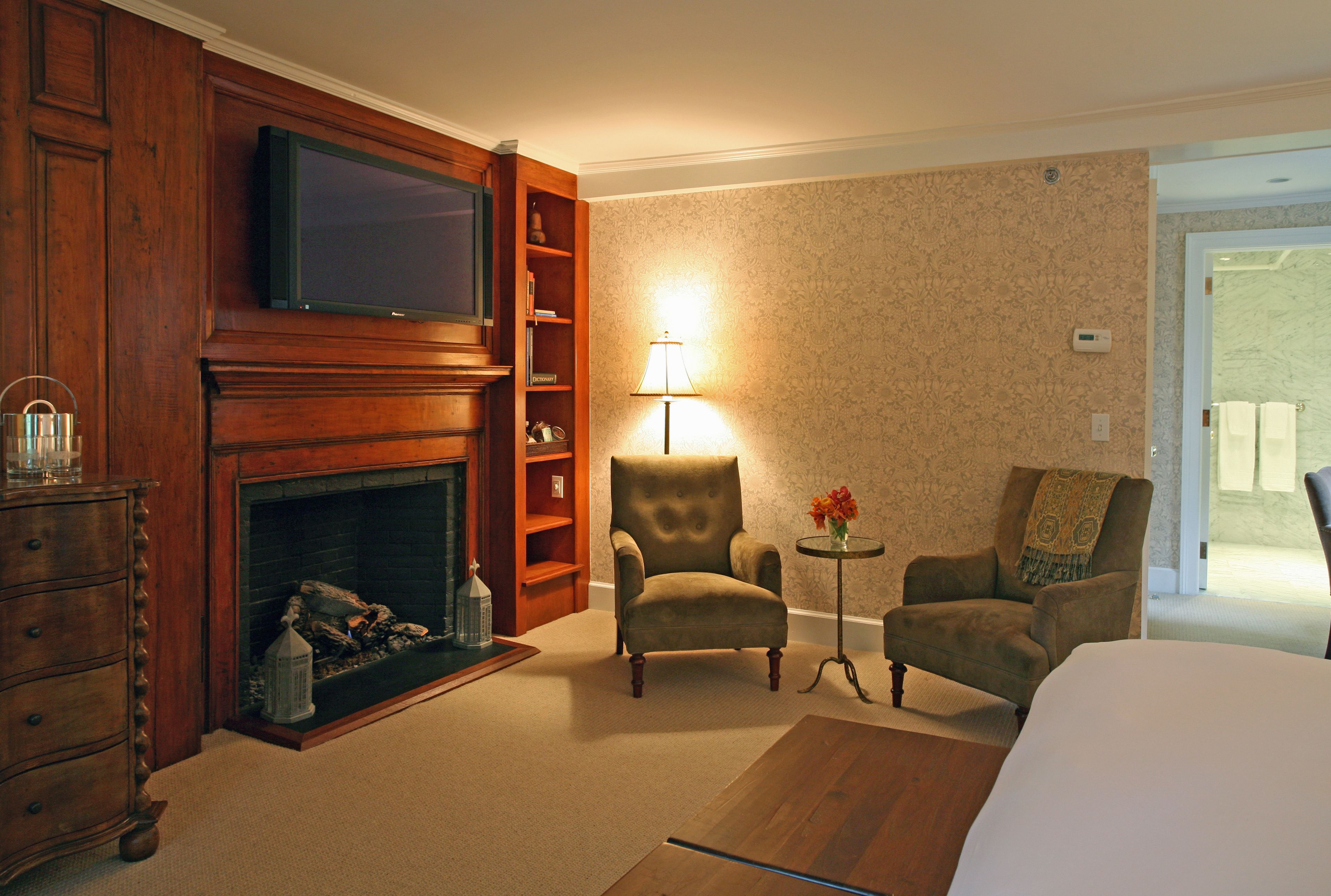 A hotel room with a fireplace, a television, two armchairs, a small table with a lamp, and a bed.