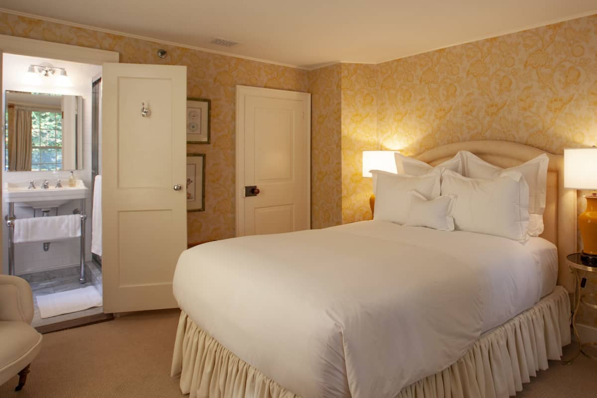A neatly arranged bedroom with a large bed, a bedside lamp, and a bathroom visible through an open door.