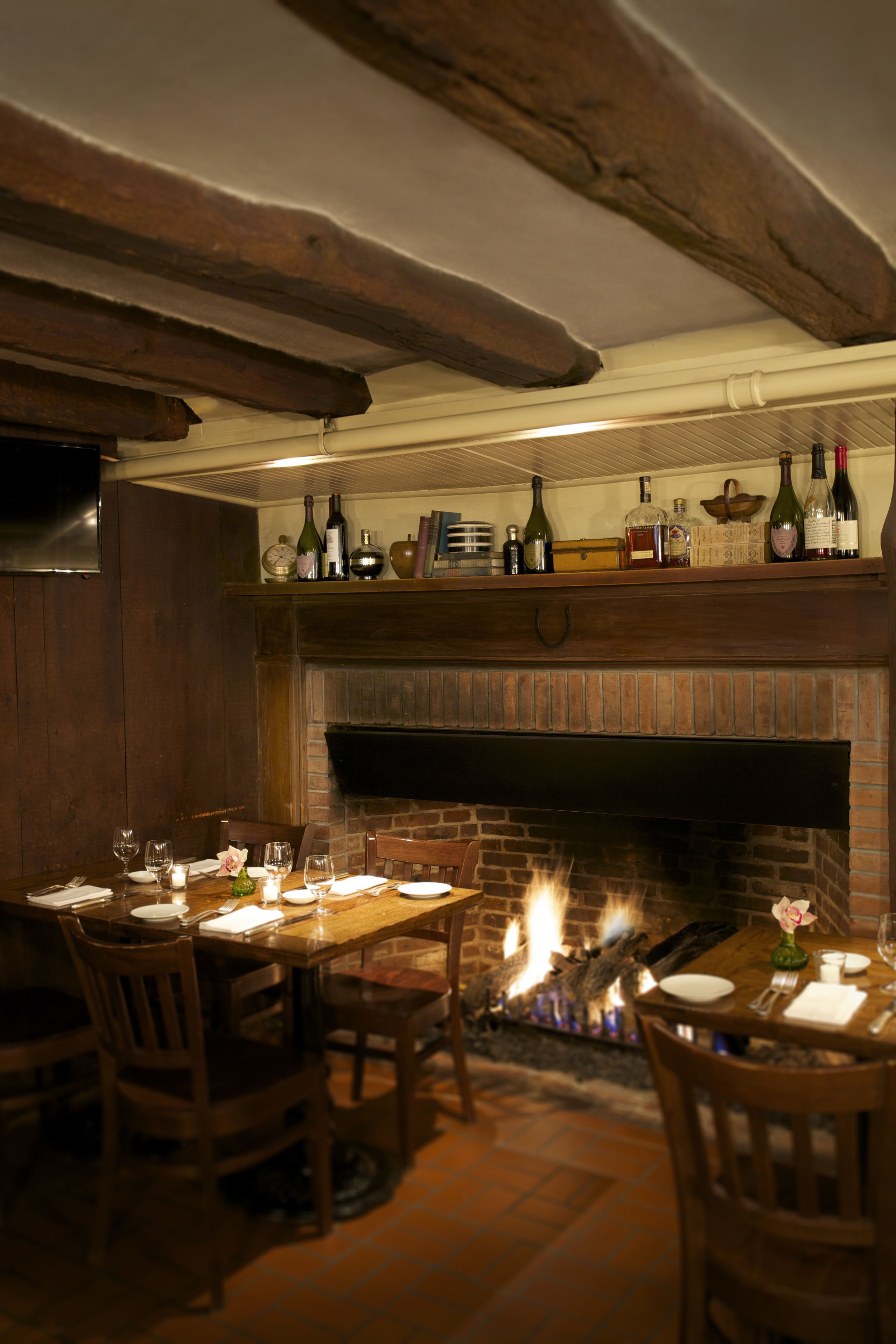 A dining area with a brick fireplace, wooden beams, and a shelf with wine bottles.