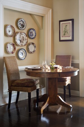 A dining area with a round wooden table, two wicker chairs, and a wall adorned with decorative plates and a framed botanical illustration.