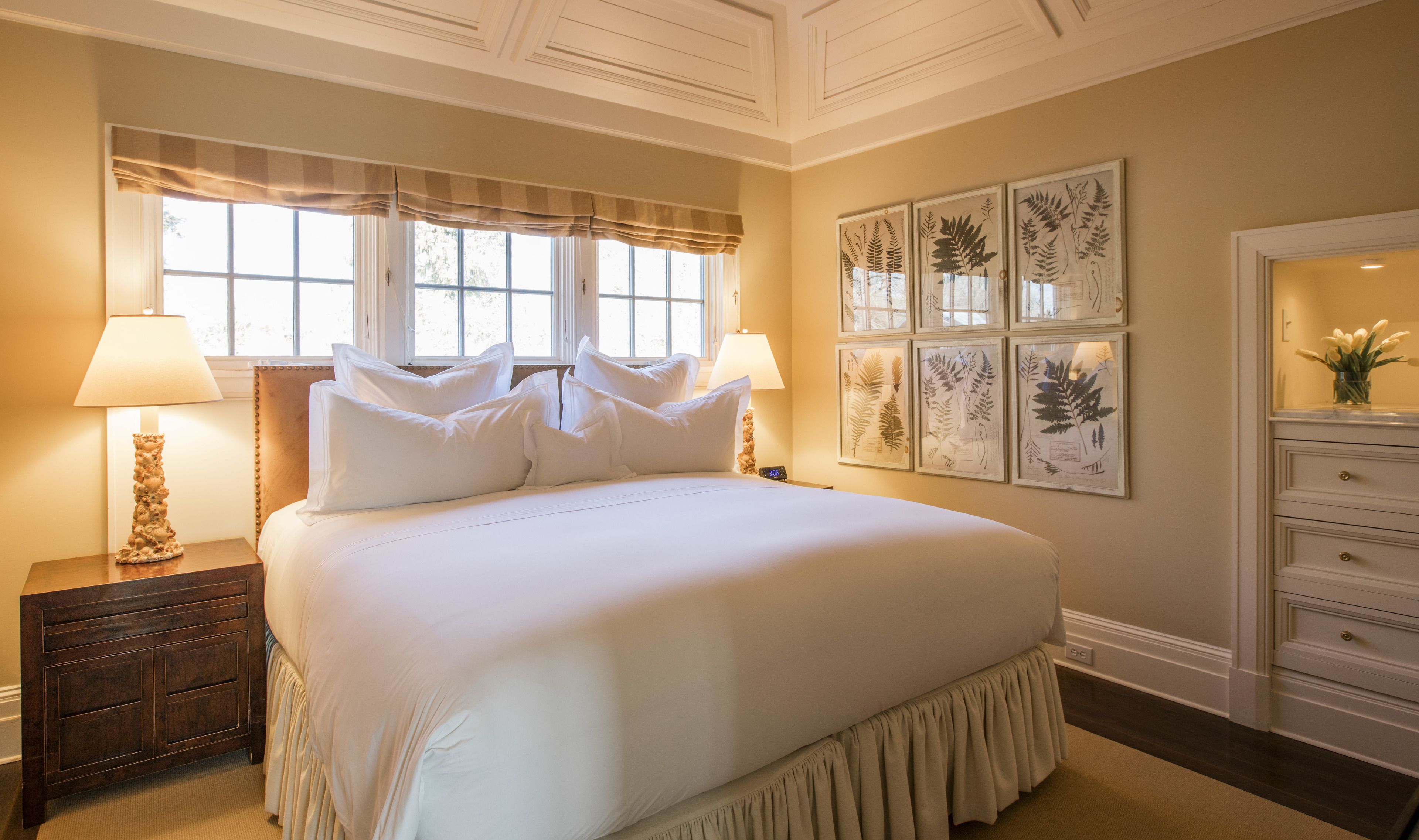 A neatly arranged bedroom with a large bed, two bedside lamps, a nightstand, a dresser, and framed botanical prints on the wall.