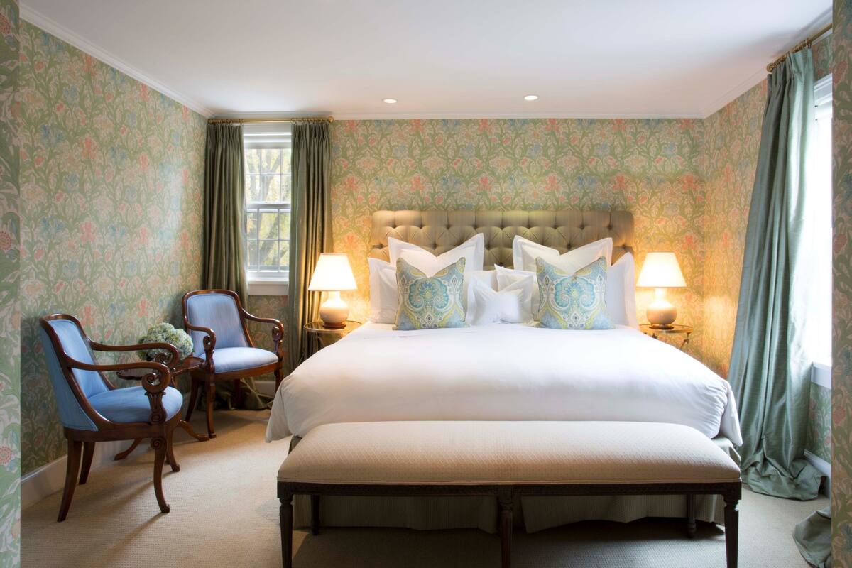 A large bed with a tufted headboard, two bedside lamps, and floral wallpaper.