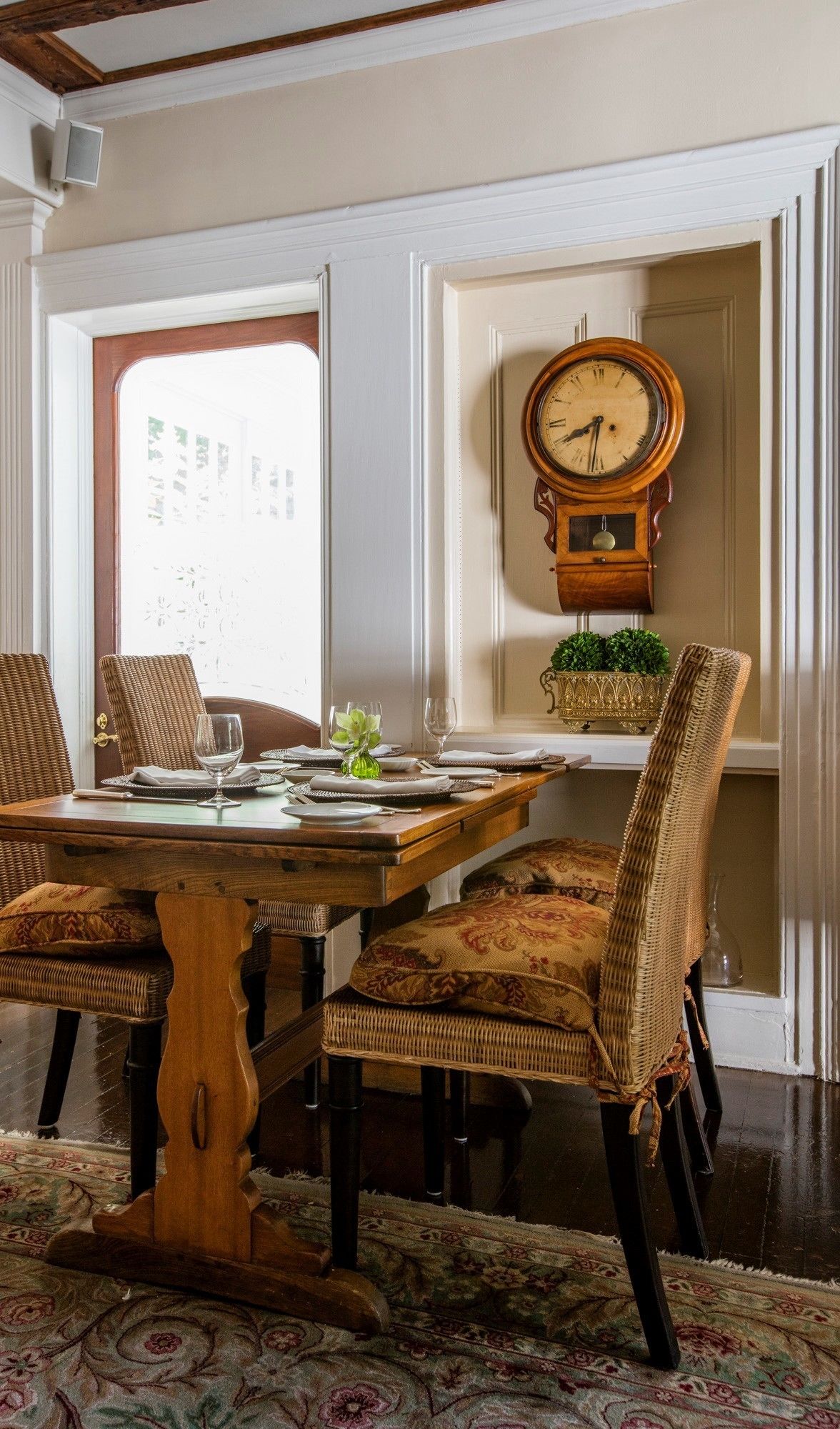A dining room with a wooden table set for four, surrounded by wicker chairs. A grandfather clock is mounted on the wall.