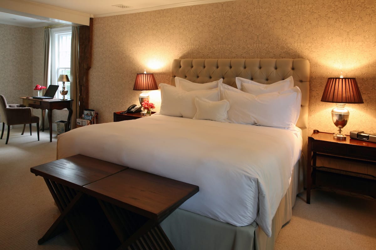 A neatly made bed with a tufted headboard, a bedside table with a lamp, and a small desk with a chair in a hotel room.
