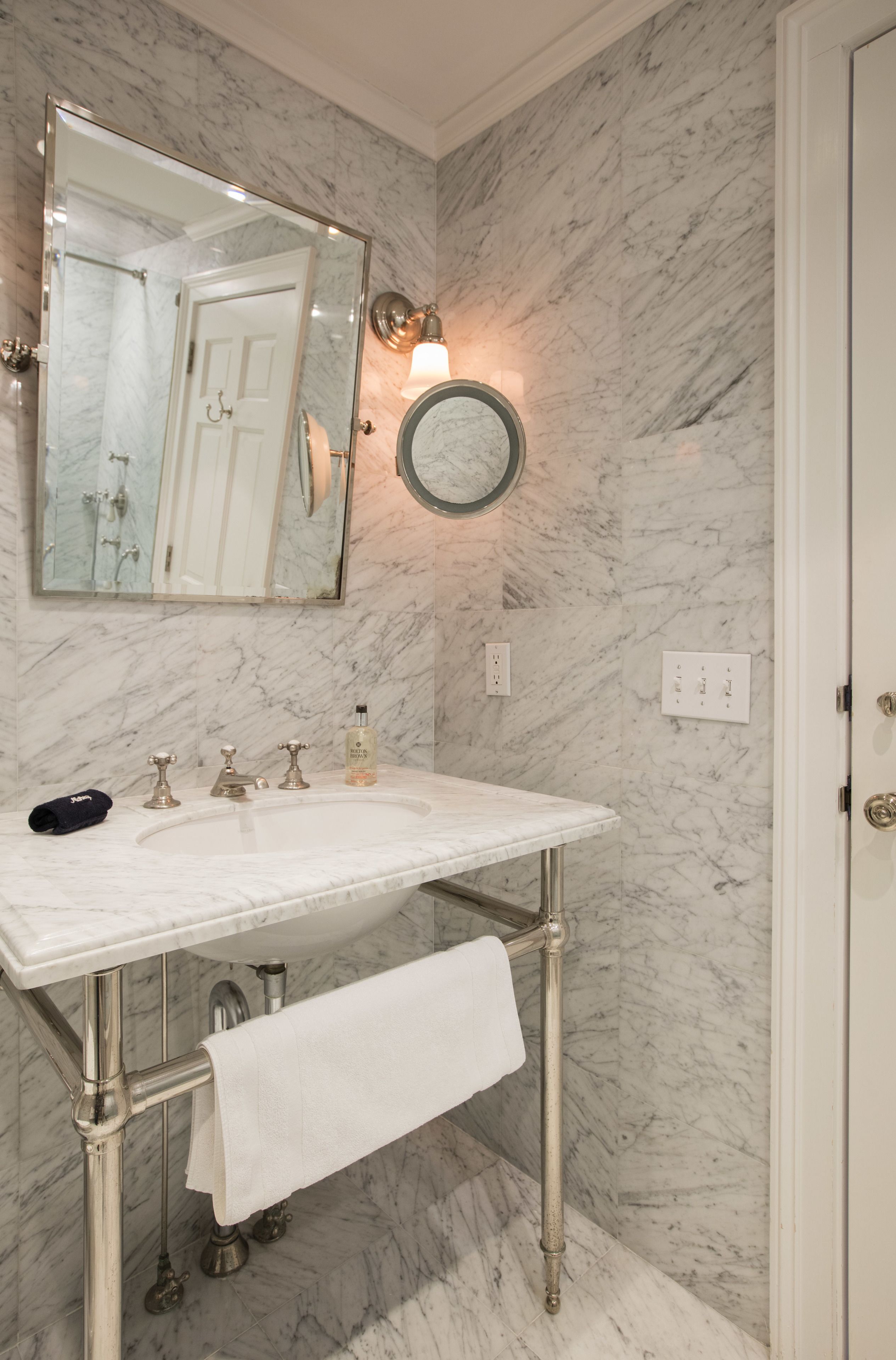 A bathroom with a marble wall, a large mirror, a sink with a towel, and a round wall-mounted light.