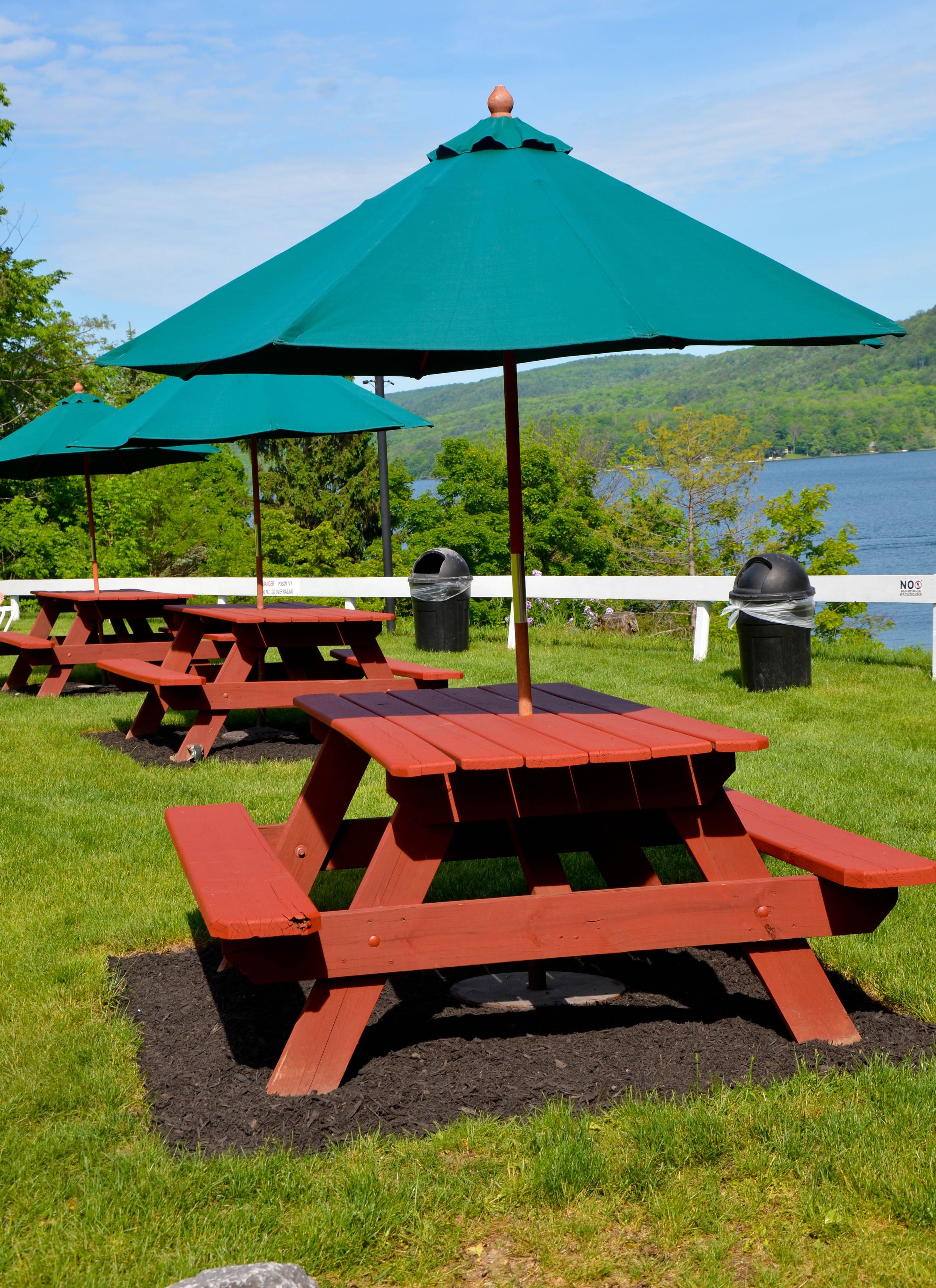 Outdoor picnic tables with green umbrellas, a white fence, and a lake in the background.