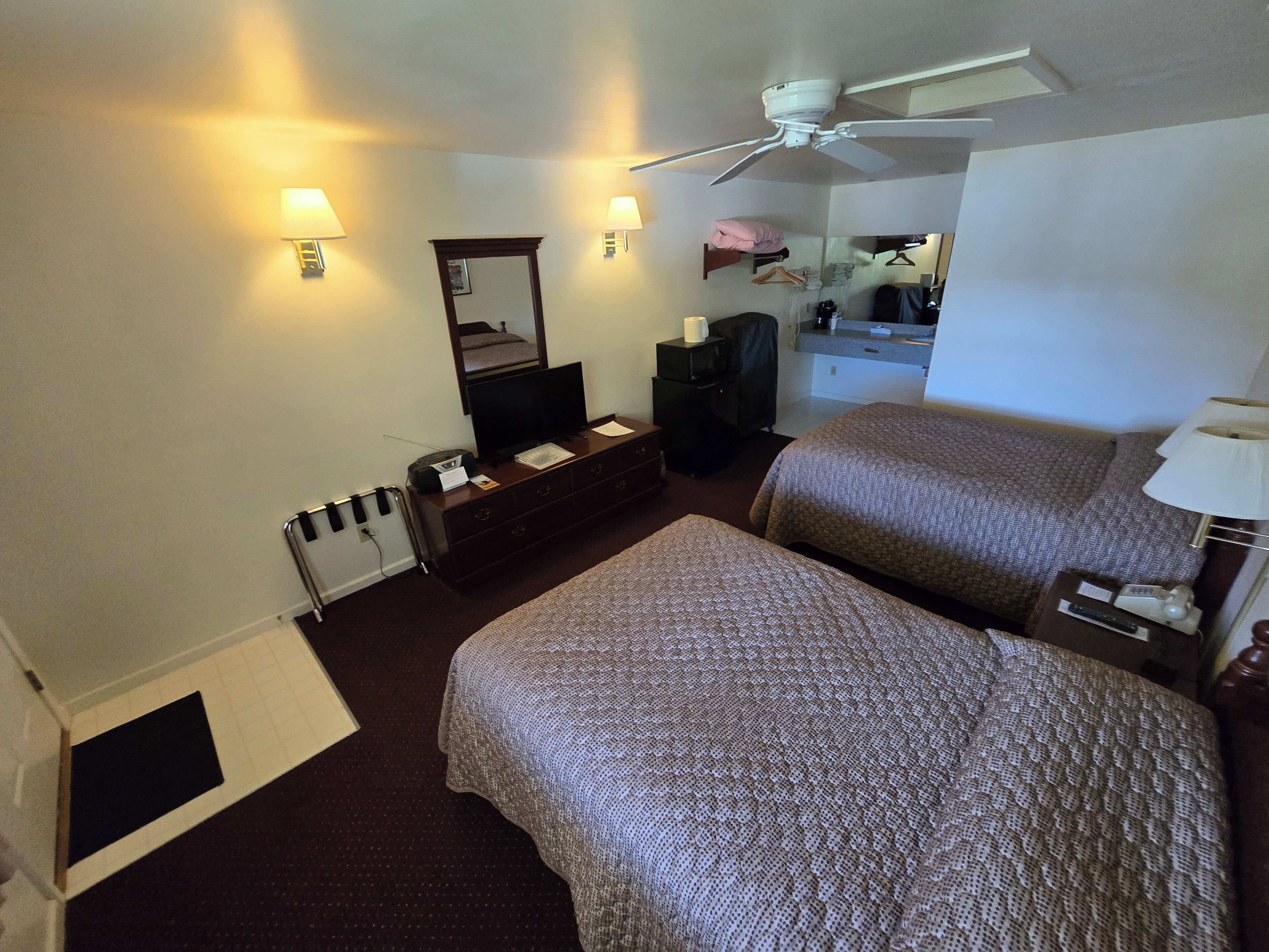 A hotel room with two beds, a TV, a dresser, a mirror, and a ceiling fan.