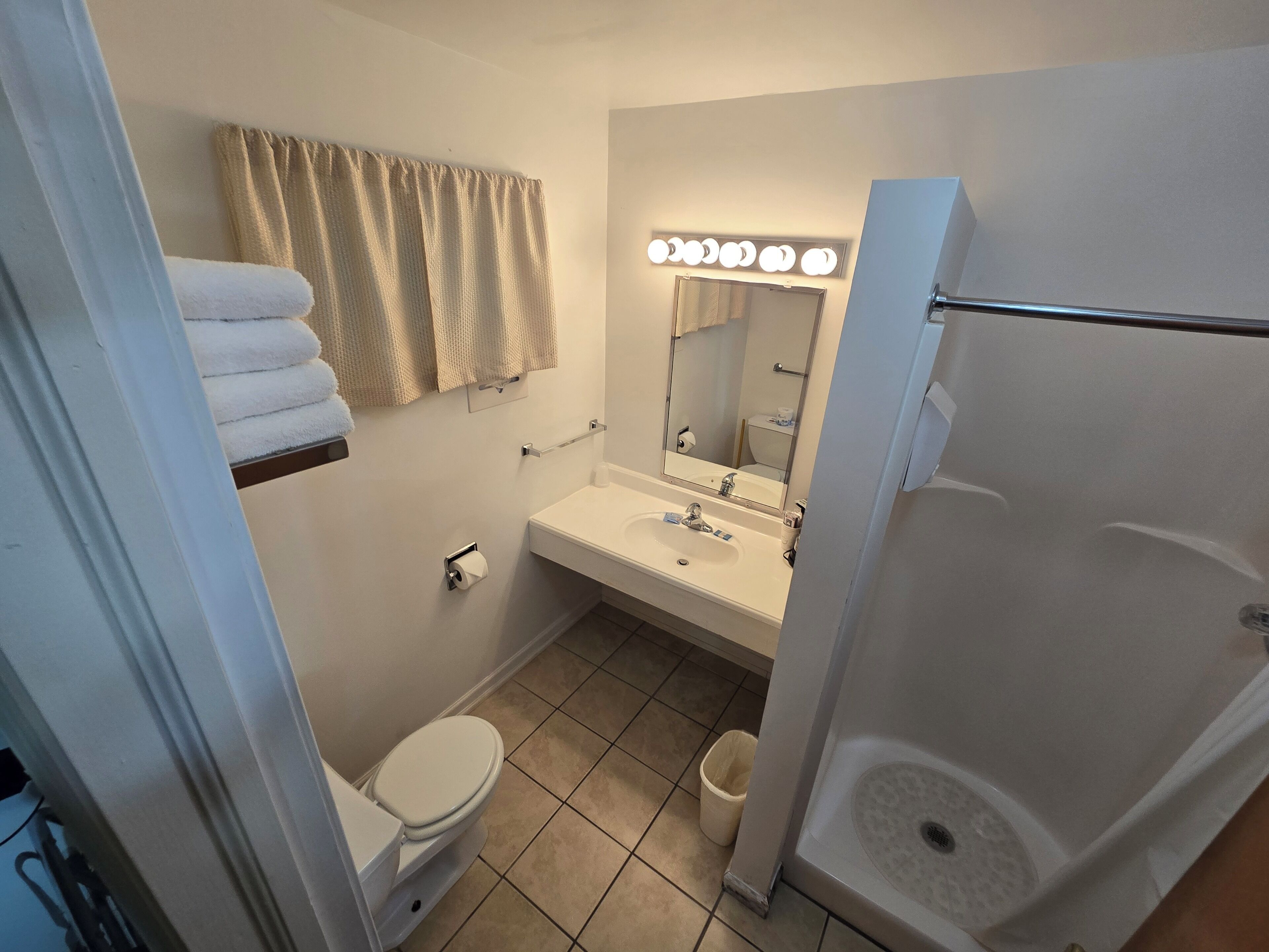 A bathroom with a toilet, sink, and shower. There are towels on a shelf, a mirror with lights, and a trash can.