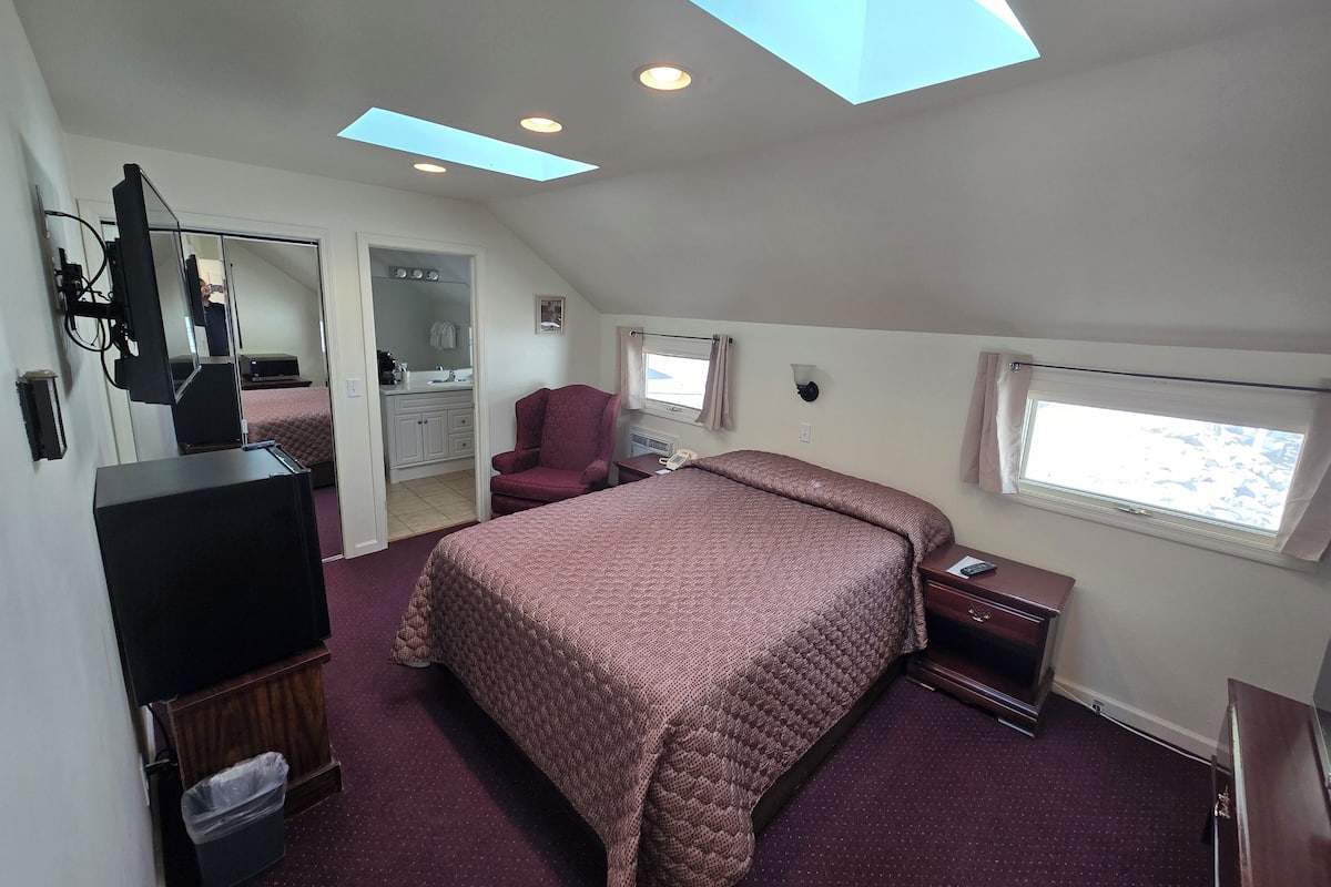 A bedroom with a large bed, a desk, a chair, a TV, a mirror, and a skylight.