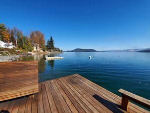 A wooden dock extending into a calm lake with clear blue water, surrounded by autumn-colored trees and houses. - Lake View Motel (Cooperstown)