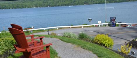 Ein roter Adirondack-Stuhl am Kiesweg entlang eines Sees, mit Blick auf das Wasser und einen bewaldeten Hügel im Hintergrund.
