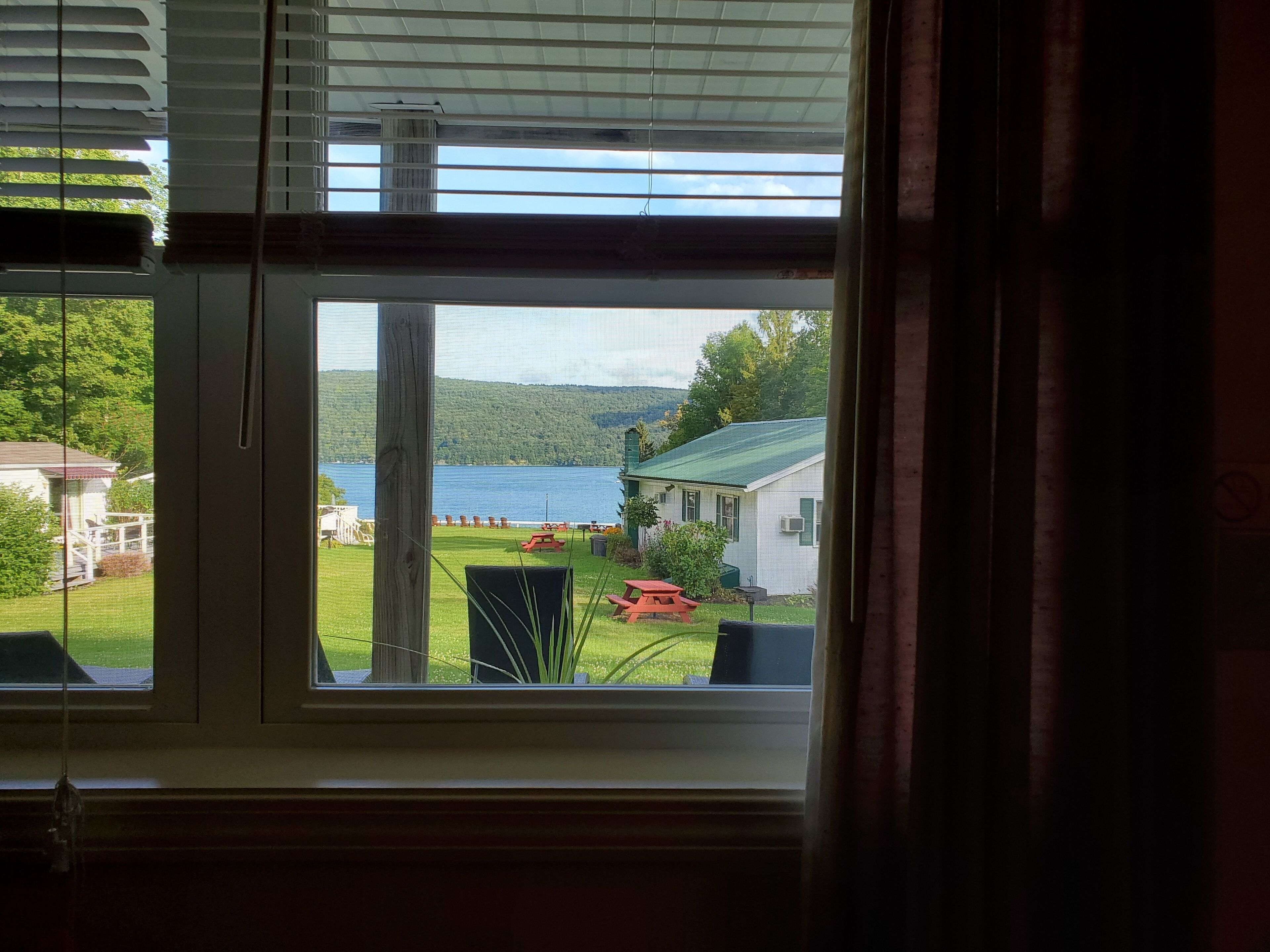 Vista a través de una ventana con persianas, sobre un área verde con una mesa de picnic y un edificio pequeño con techo verde.