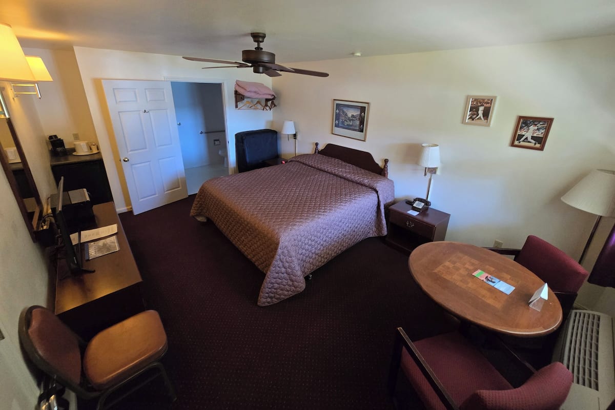 A hotel room with a bed, a desk with a chair, a small table with a chair, a ceiling fan, and a door leading to a bathroom.