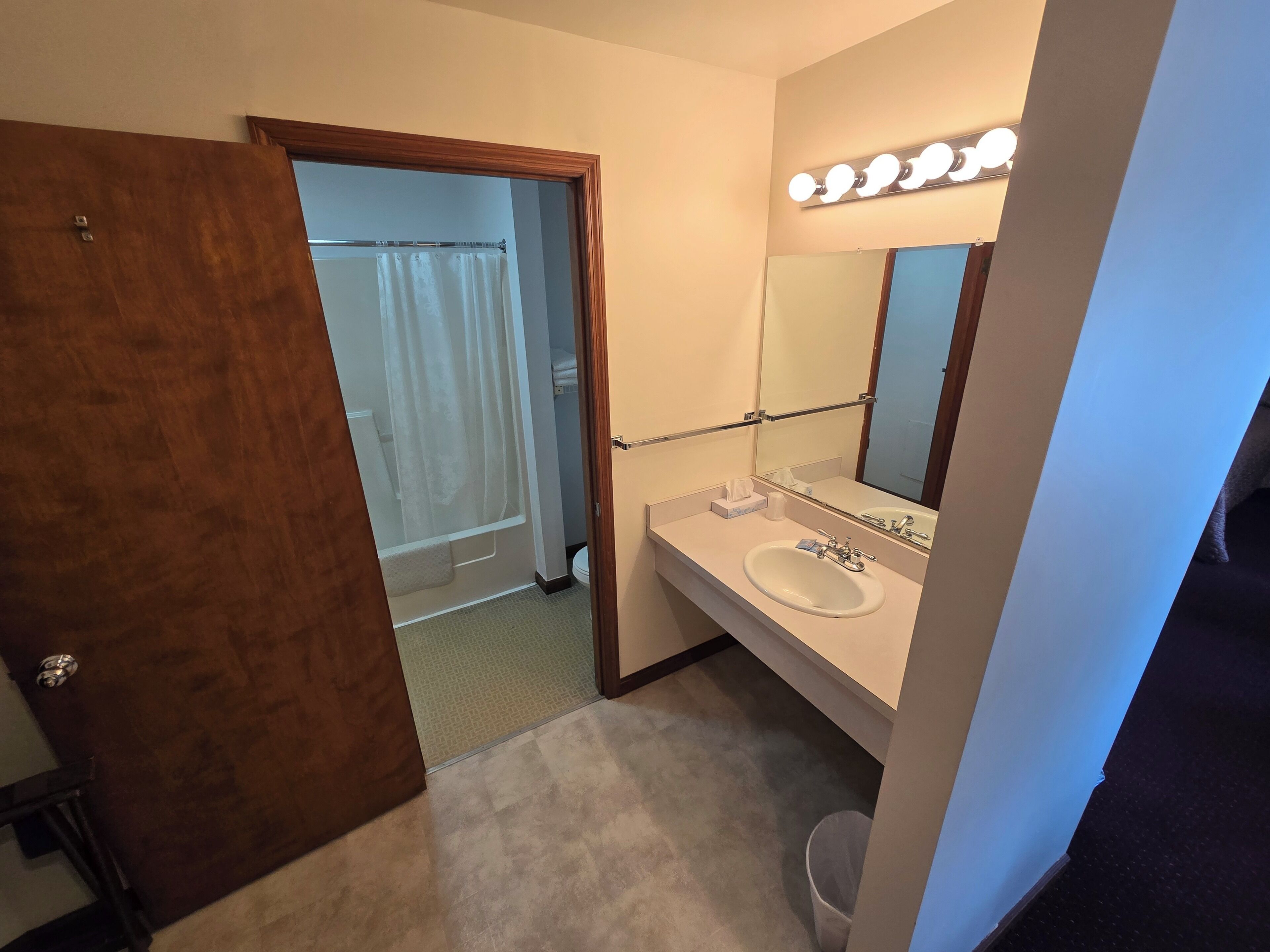 Un baño con un lavamanos blanco, un espejo con iluminación, un perchero para toallas y una cortina de ducha.