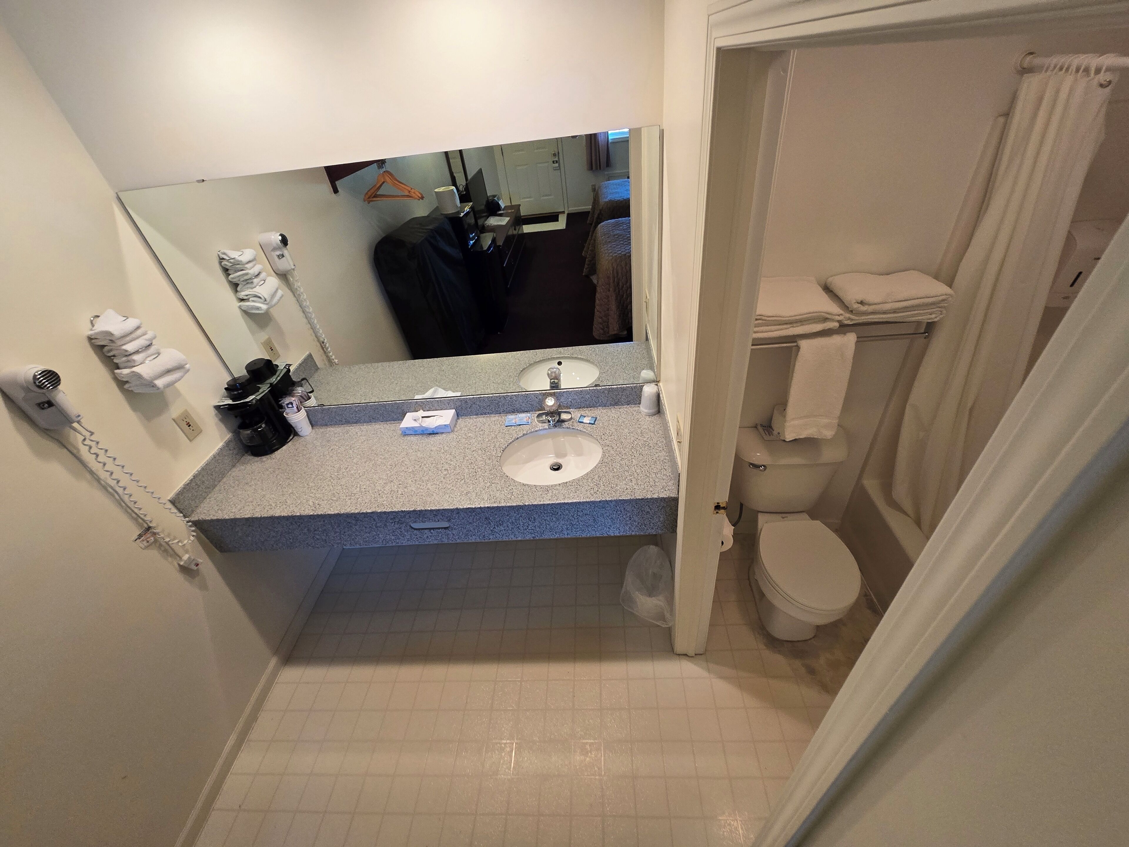 Ein Badezimmer mit Waschbecken, Spiegel und Toilette. Ein Fön ist an der Wand angebracht und ein Handtuchhalter mit Handtüchern vorhanden.
