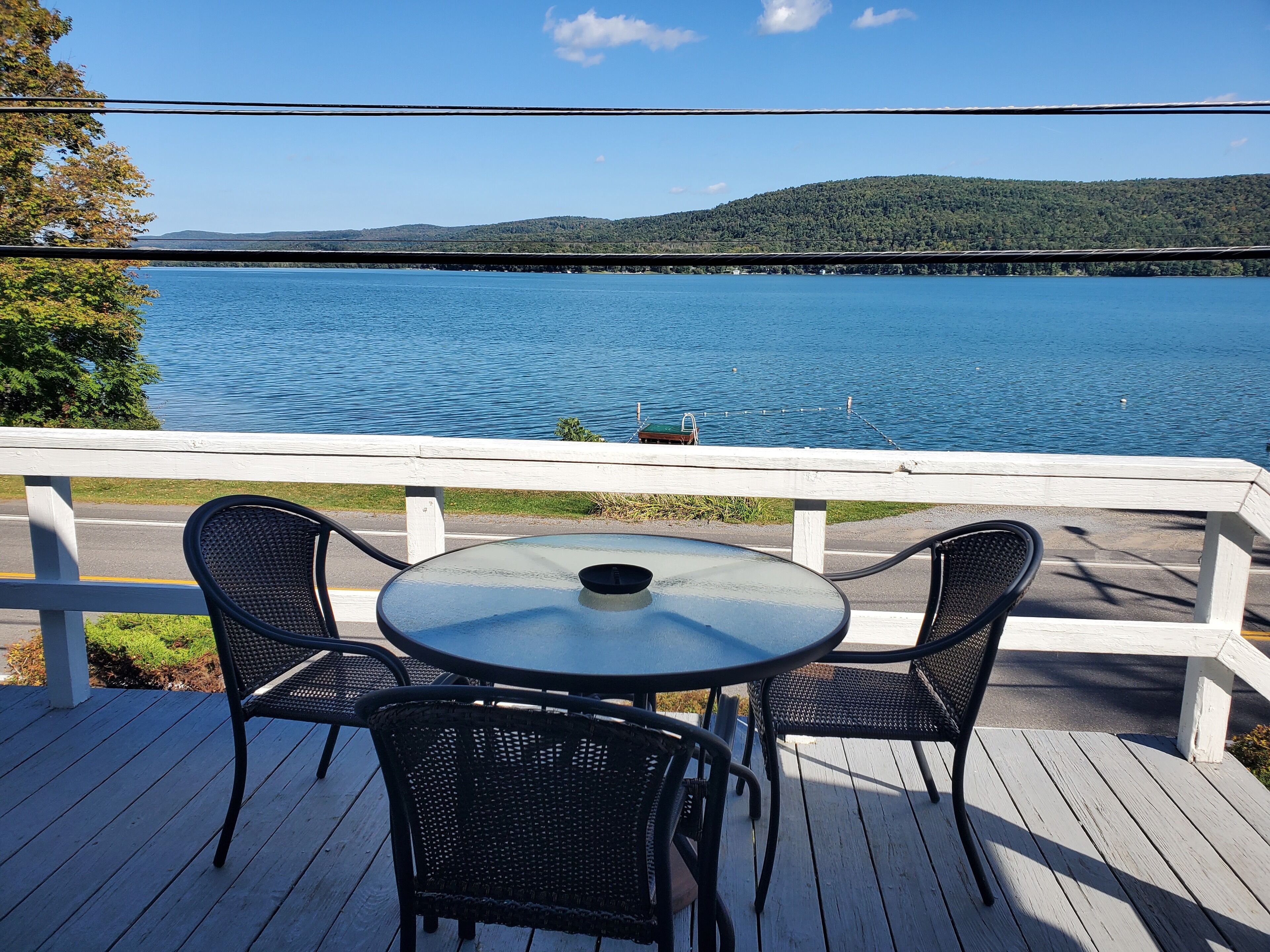 Una terraza con una mesa redonda y dos sillas, con vista a un lago que incluye un puente y colinas al fondo.