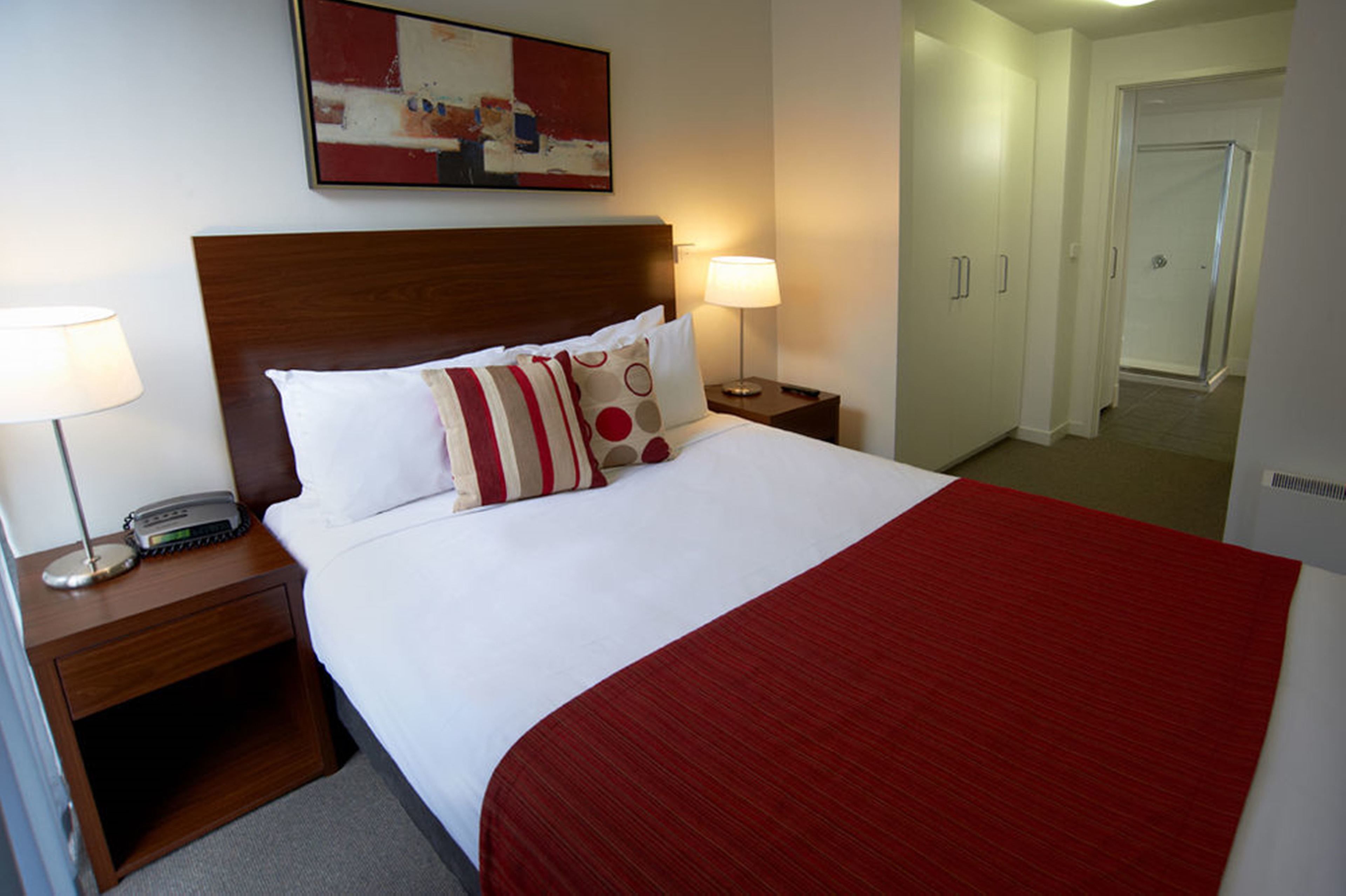 A hotel room with a large bed, two bedside tables, a nightstand, a red throw blanket, and a painting on the wall.