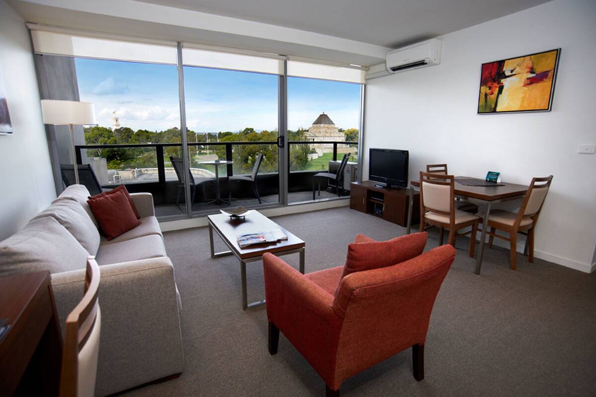 A modern living room with a sofa, red armchair, and a dining table with chairs. A television is on a stand, and there's a large window offering a view of a building and greenery.