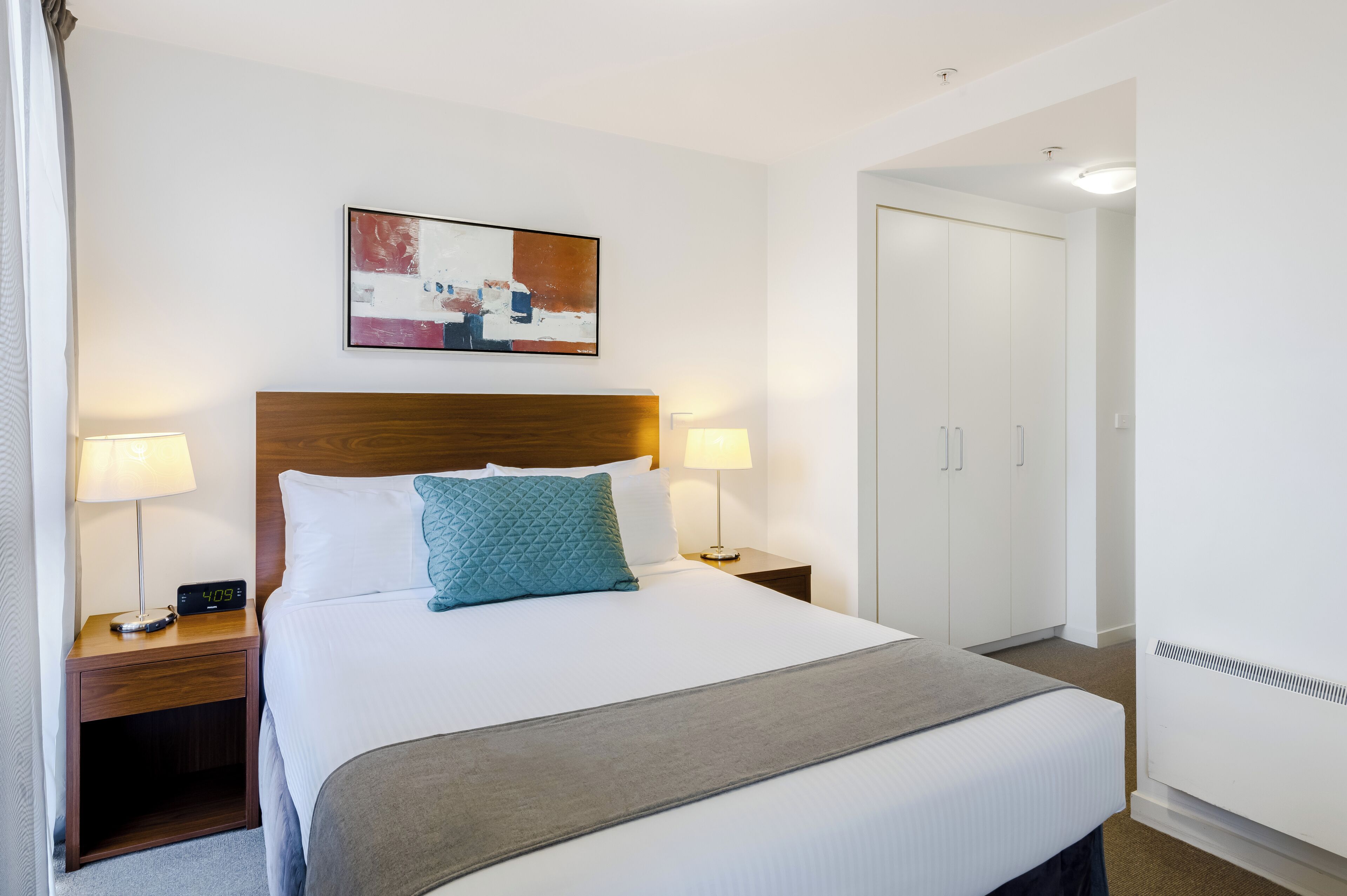 Una habitación de hotel con una cama, dos lámparas de noche, un reloj digital, un cuadro, un armario y un calentador.