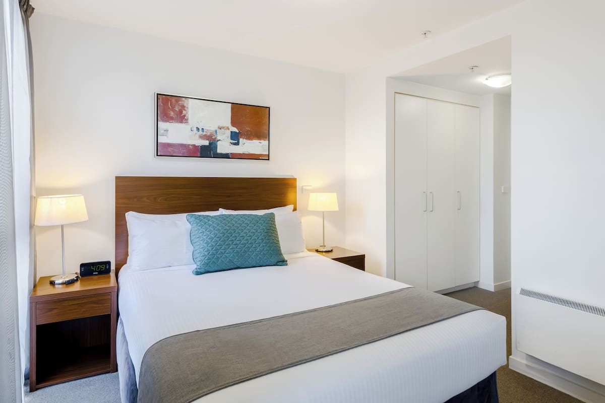 A hotel room with a bed, two bedside lamps, a digital clock, a painting, a wardrobe, and a heater.