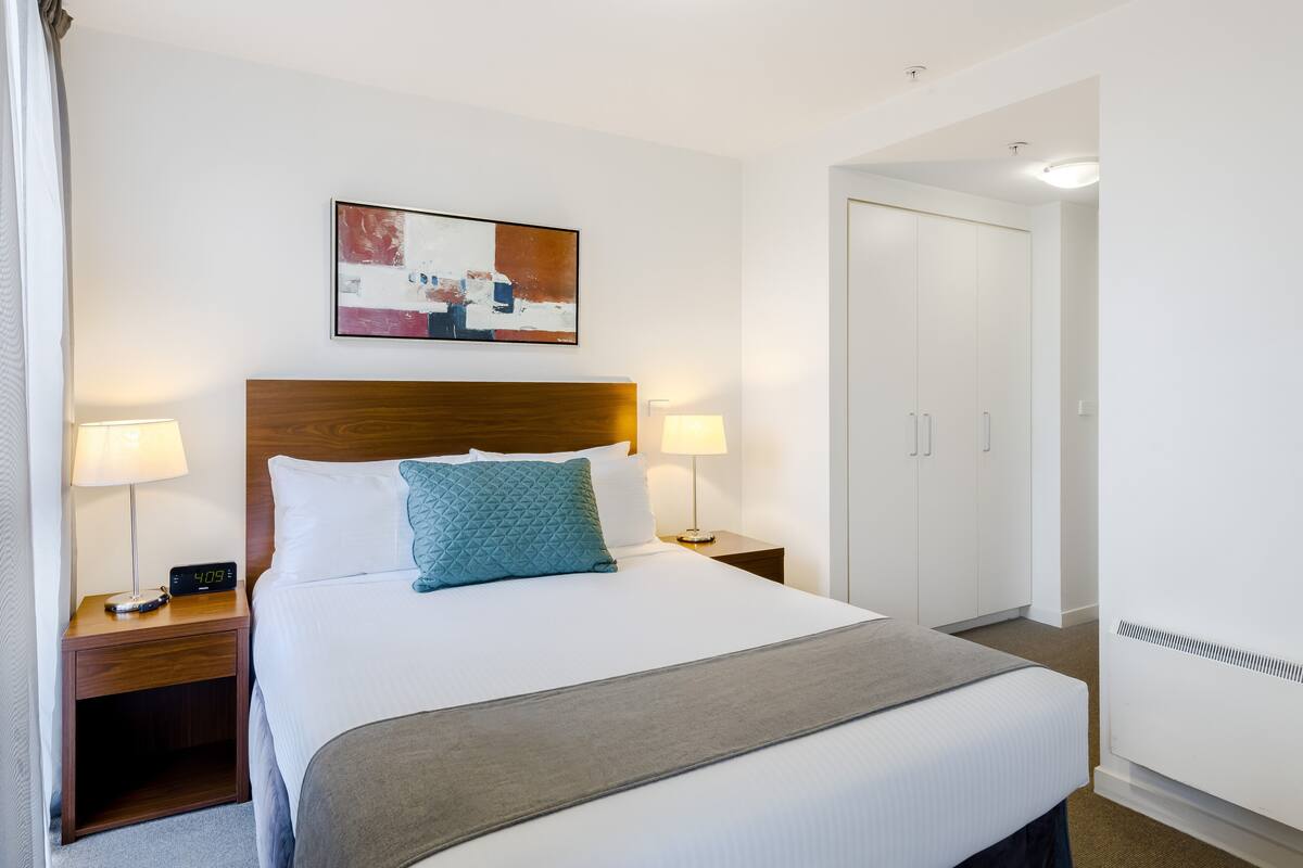 A hotel room with a bed, two bedside lamps, a digital clock, a painting, a wardrobe, and a heater.