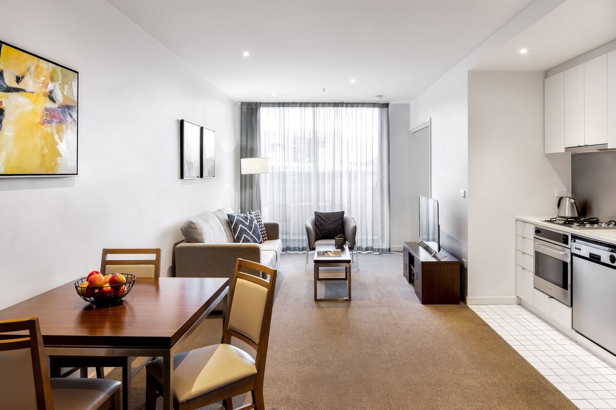 A modern living room with a dining table, chairs, a sofa, a TV, and a kitchen area with appliances.