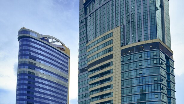 Dos edificios modernos de gran altura con fachadas de vidrio reflectante, uno de diseño curvo y el otro con forma rectangular y recta.