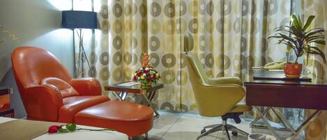 A hotel room with a bed, an orange chair, a yellow chair, a small table with a vase of flowers, and a desk with a plant.