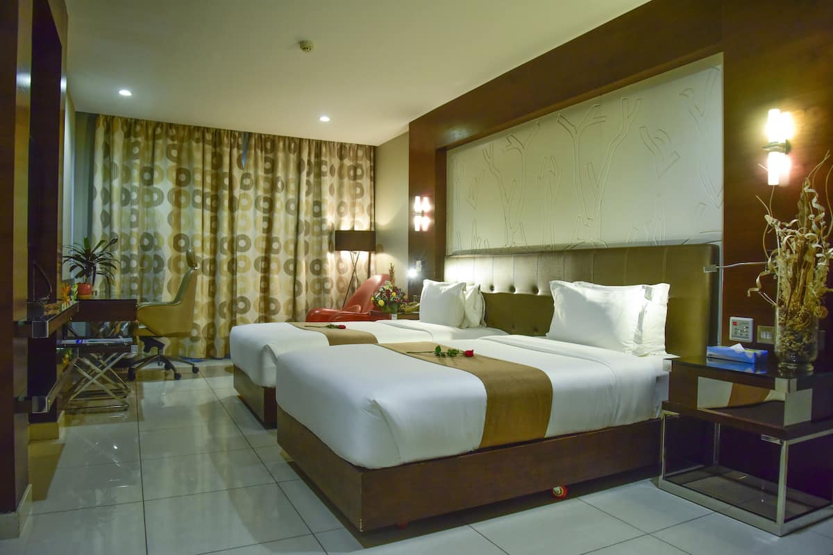 A hotel room with a large bed, two bedside tables, a desk with a chair, a potted plant, and a decorative wall panel.