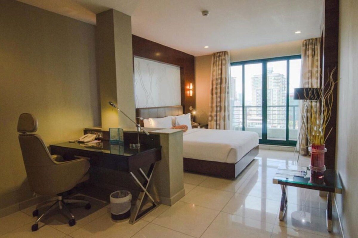 Habitación de hotel con cama, escritorio, silla y un amplio ventanal con vistas a la ciudad.
