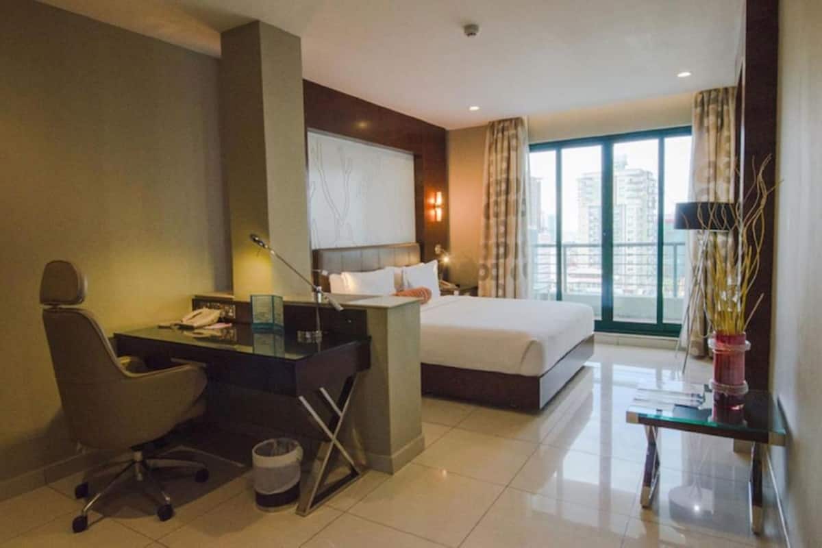 A hotel room with a bed, desk, chair, and a large window with a city view.