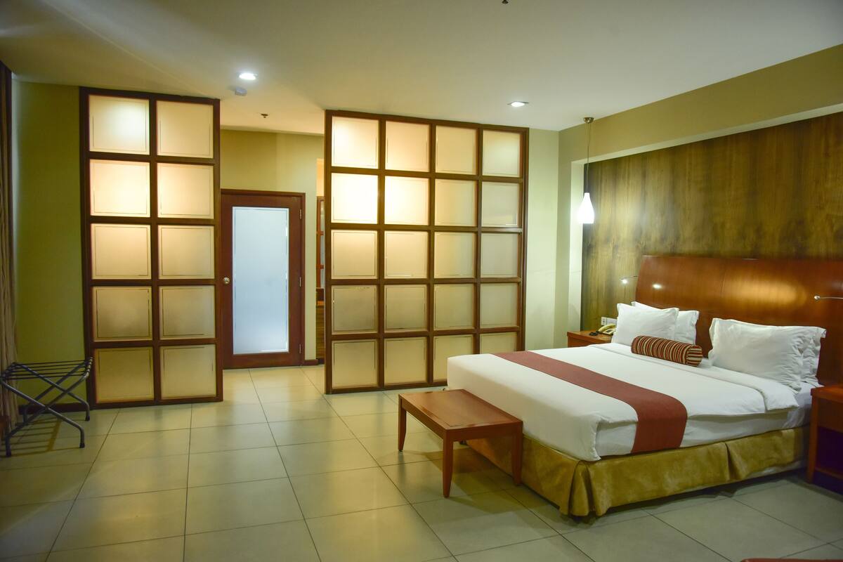 A hotel room with a large bed, a wooden headboard, a bench, a wardrobe with sliding doors, and a small table.