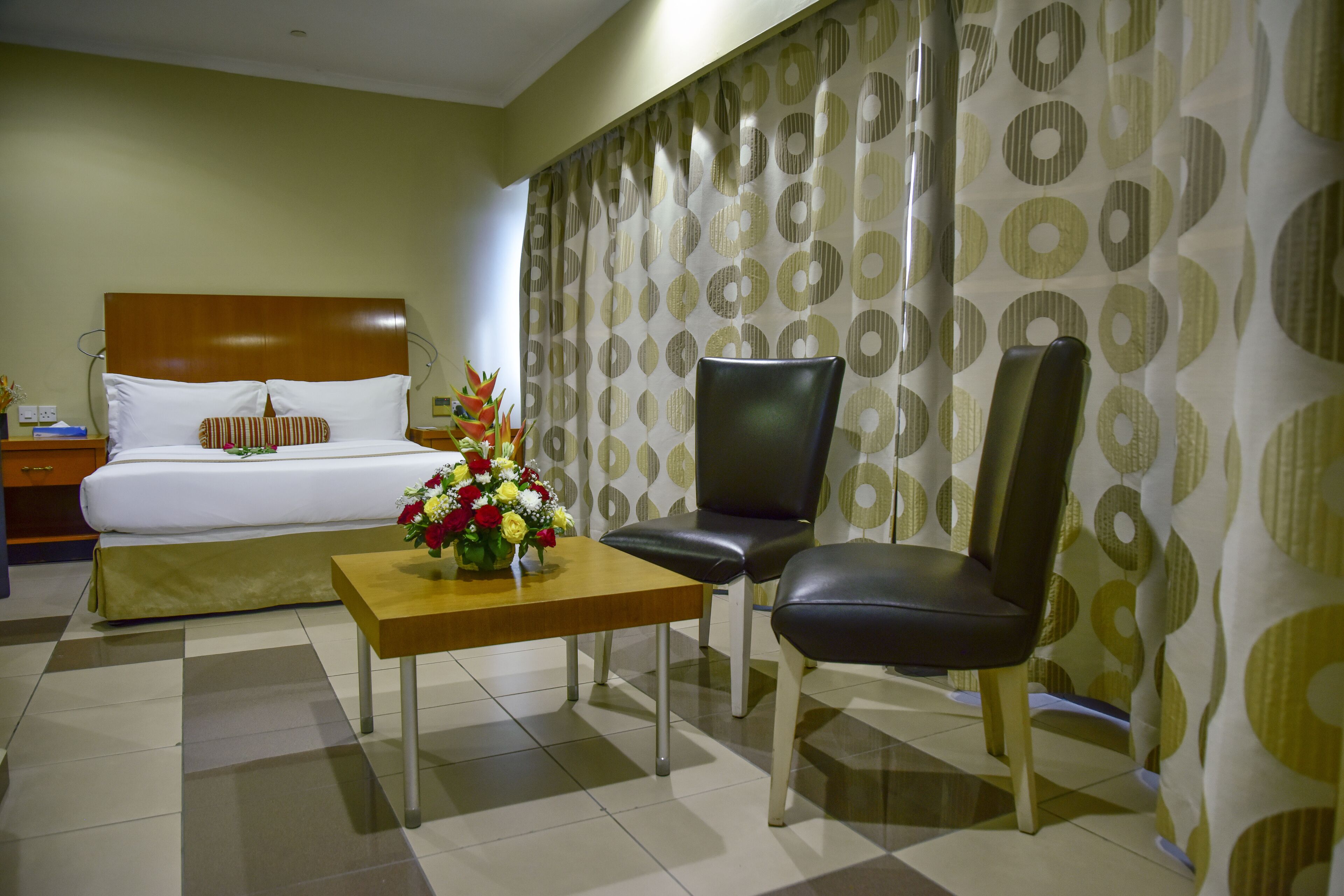 Habitación de hotel con una cama, una mesa de madera con un florero, dos sillones de cuero y una cortina estampada.