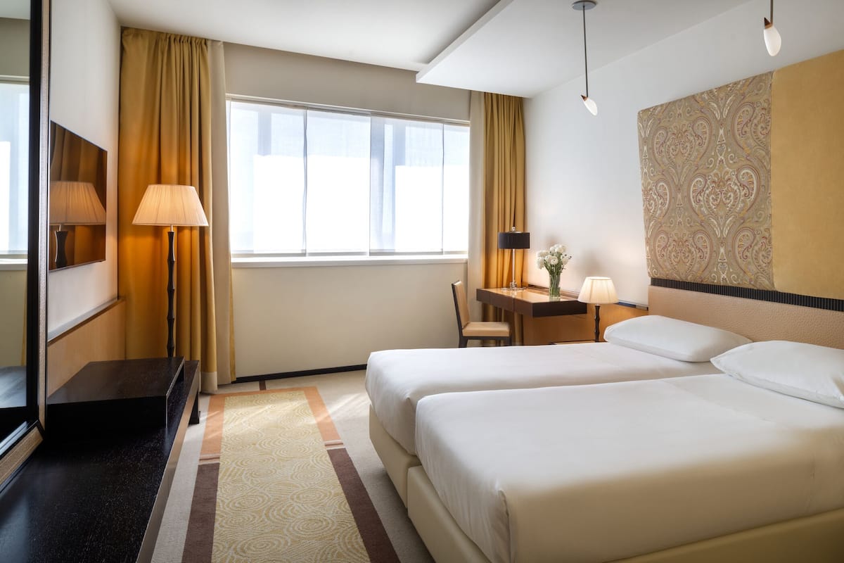 Habitación de hotel con dos camas, un escritorio con silla, una lámpara y un ventanal con cortinas.