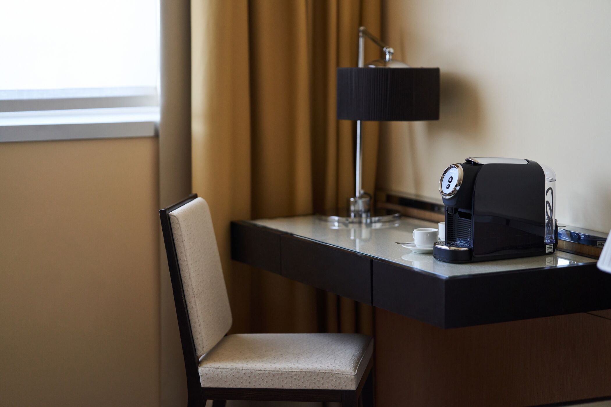 A hotel room with a desk, a chair, a coffee maker, and a lamp.