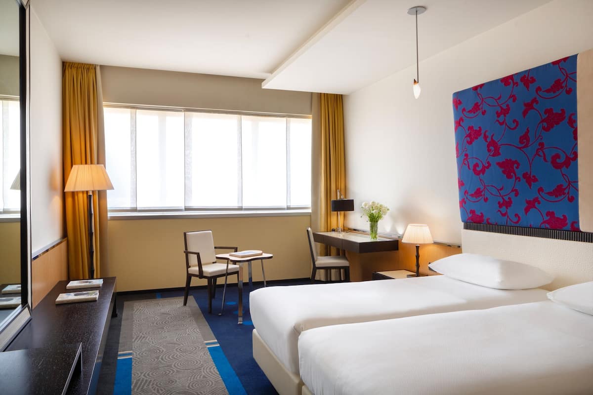 Habitación de hotel con dos camas, un escritorio, una silla, una lámpara y un ventanal grande con cortinas.