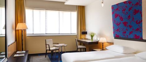 A hotel room with two beds, a desk, a chair, a lamp, and a large window with curtains.