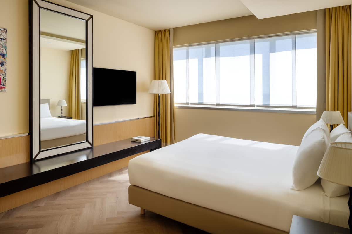 Habitación de hotel con una cama grande, un televisor de pantalla plana en la pared, una mesita de noche con lámpara y una ventana con cortinas.