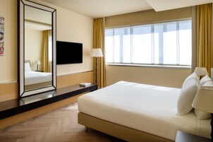 A hotel room with a large bed, a flat-screen TV mounted on the wall, a bedside table with a lamp, and a window with curtains.