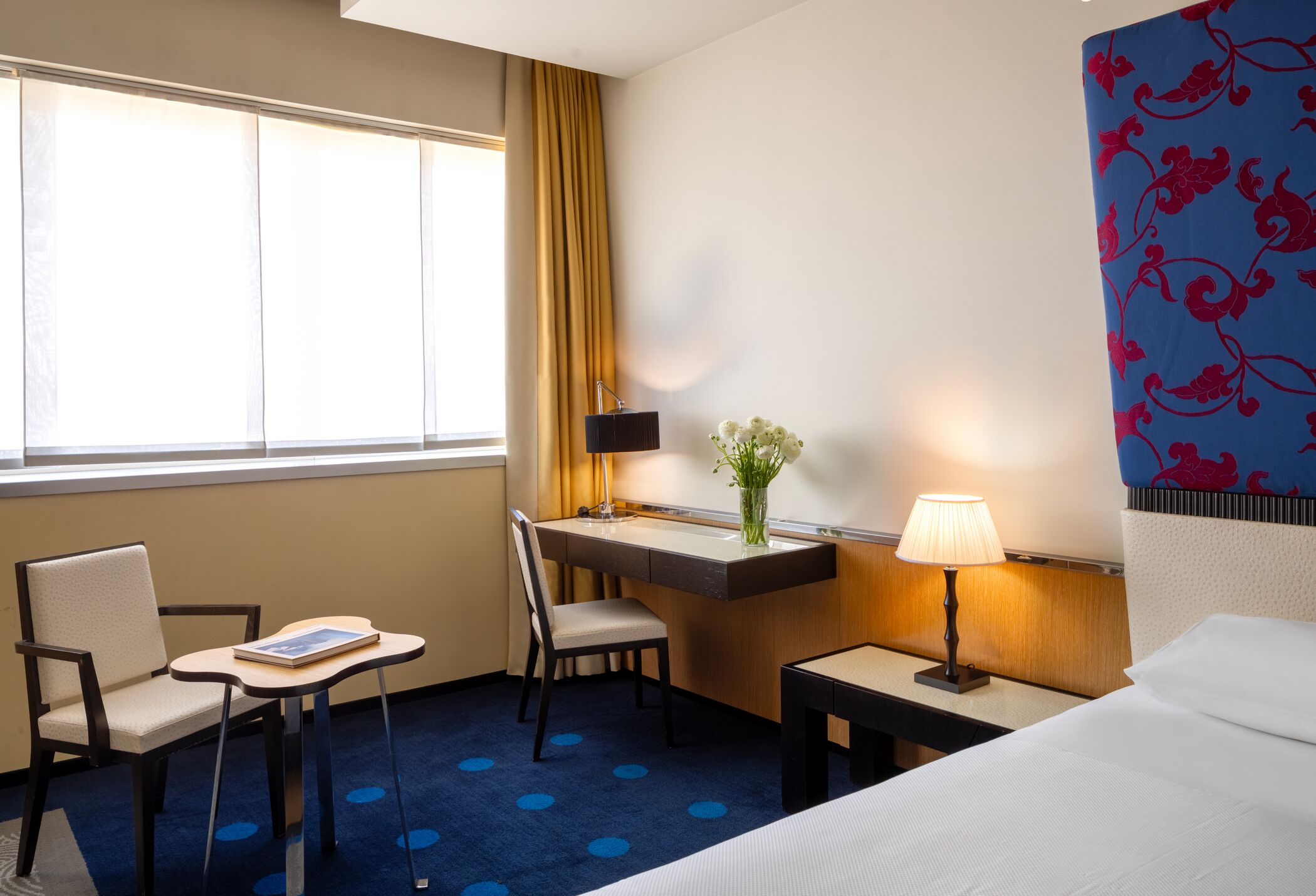 A hotel room with a bed, a desk, two chairs, a small table, a lamp, and a vase with flowers.