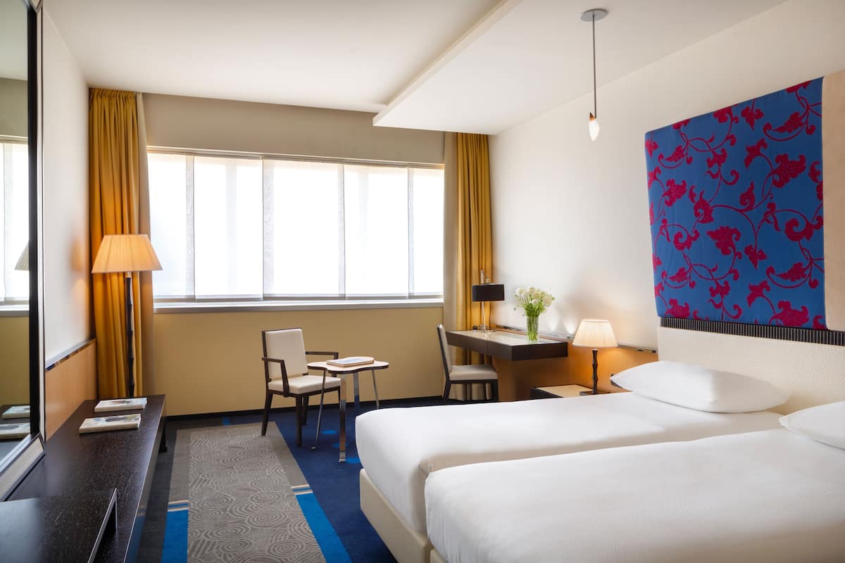 Habitación de hotel con dos camas, un escritorio, una silla, una lámpara y un ventanal con cortinas.